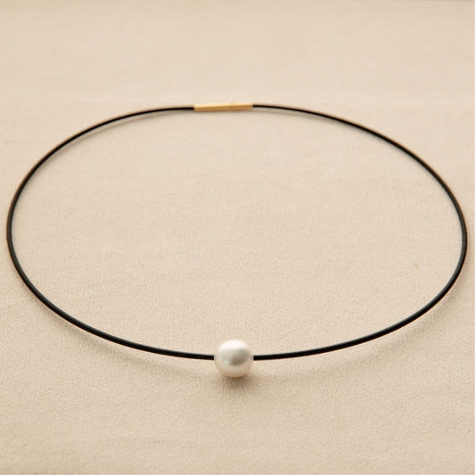 A minimalist black stainless steel necklace with a single white Australian South Sea pearl, displayed on a textured beige background.