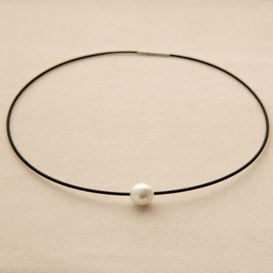 A minimalist black stainless steel necklace with a single white Australian South Sea pearl, displayed on a textured beige background.