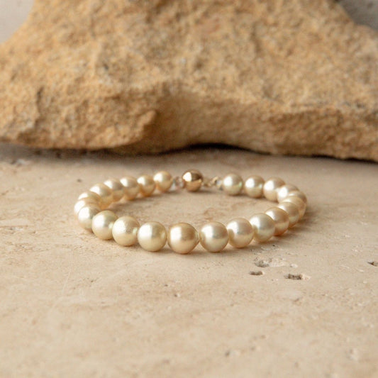 A pearl strand bracelet made of round, lustrous Australian Akoya pearls in a warm cream tone, resting on a textured stone surface with a larger rock blurred in the background.