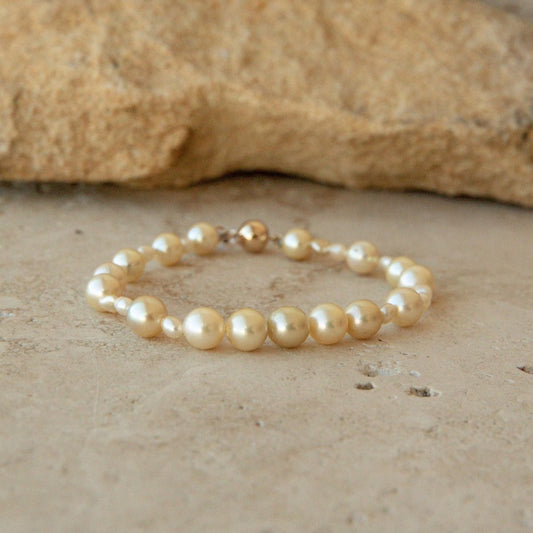 A pearls trand bracelet made of cream-colored Keshi and Akoya pearls and a small 9ct yellow gold ball clasp, resting on a textured beige stone surface with a larger rock in the background.