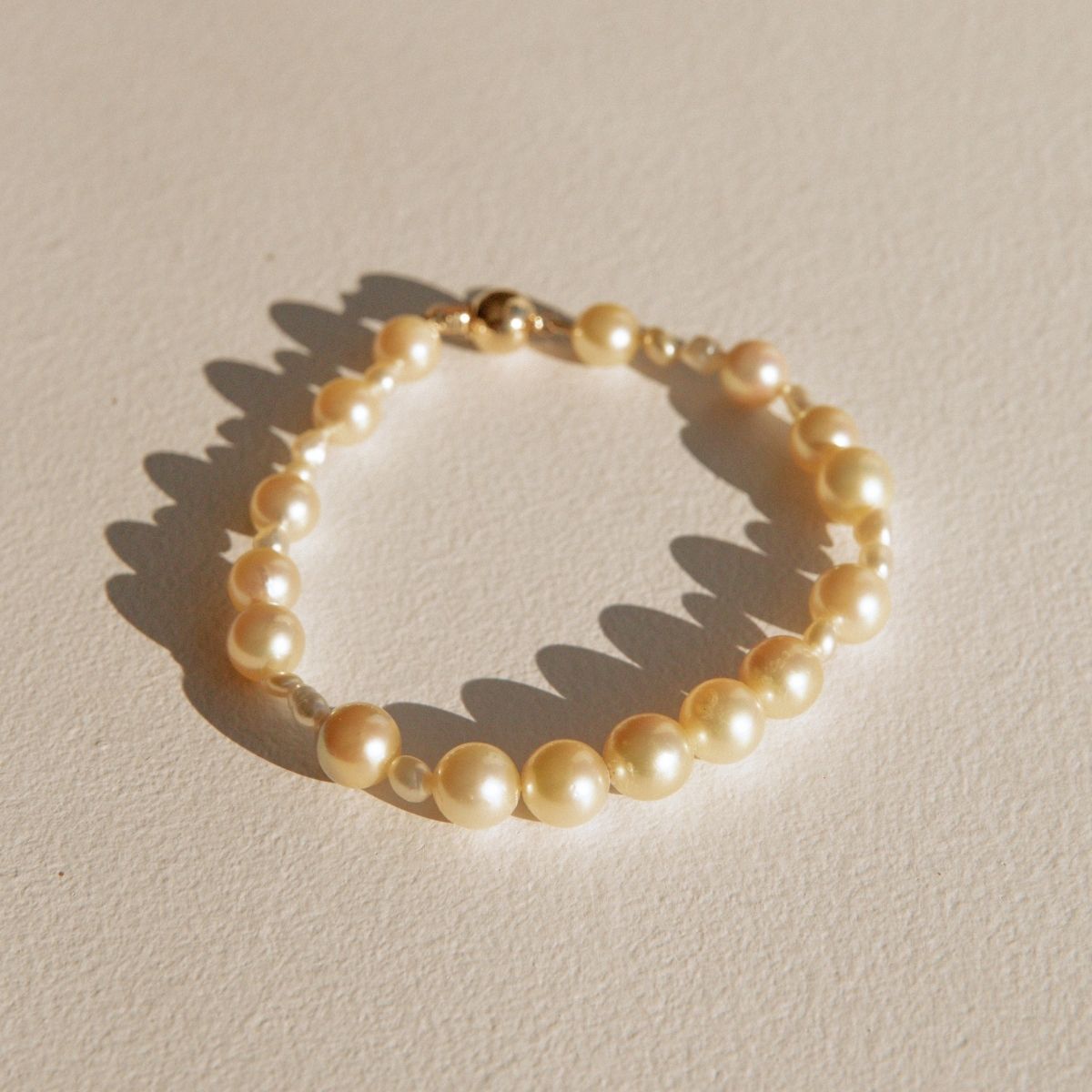 A delicate pearl strand bracelet with a gold clasp, displayed on a textured beige surface with a soft shadow.