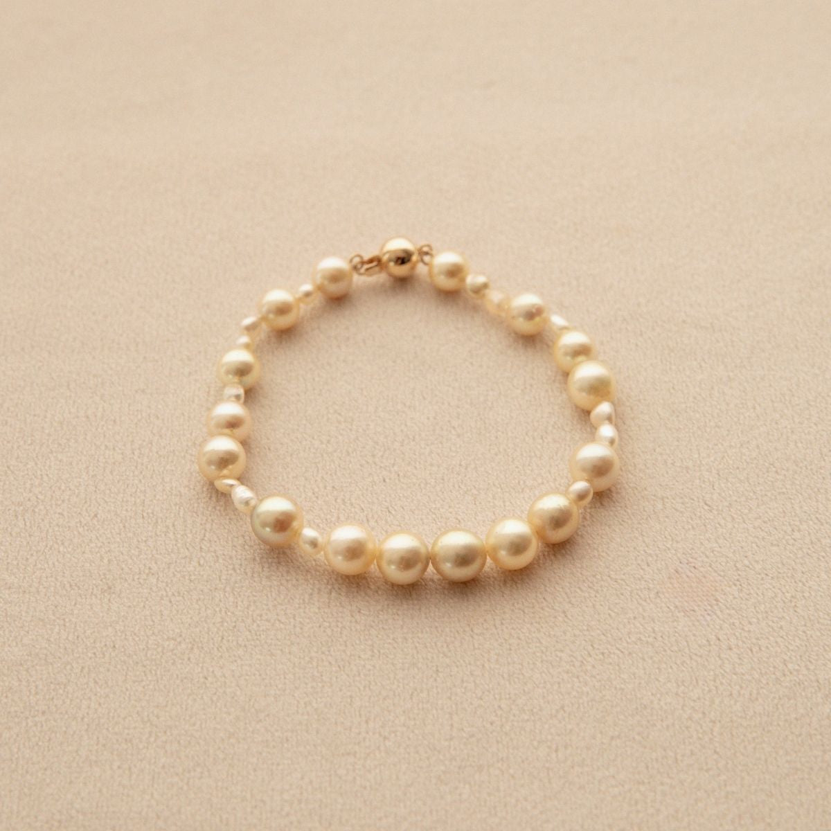 A delicate pearl strand bracelet made of small gold toned Australian Keshi and Akoya pearls with a gold clasp, displayed on a textured beige surface.