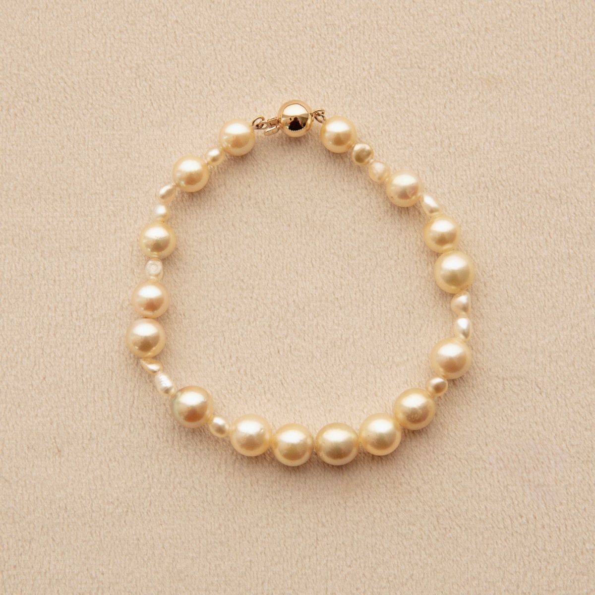 An Australian South Sea pearl strand bracelet made of golden Keshi and Akoya pearls of varying sizes, accented with a gold clasp, displayed on a textured beige background.