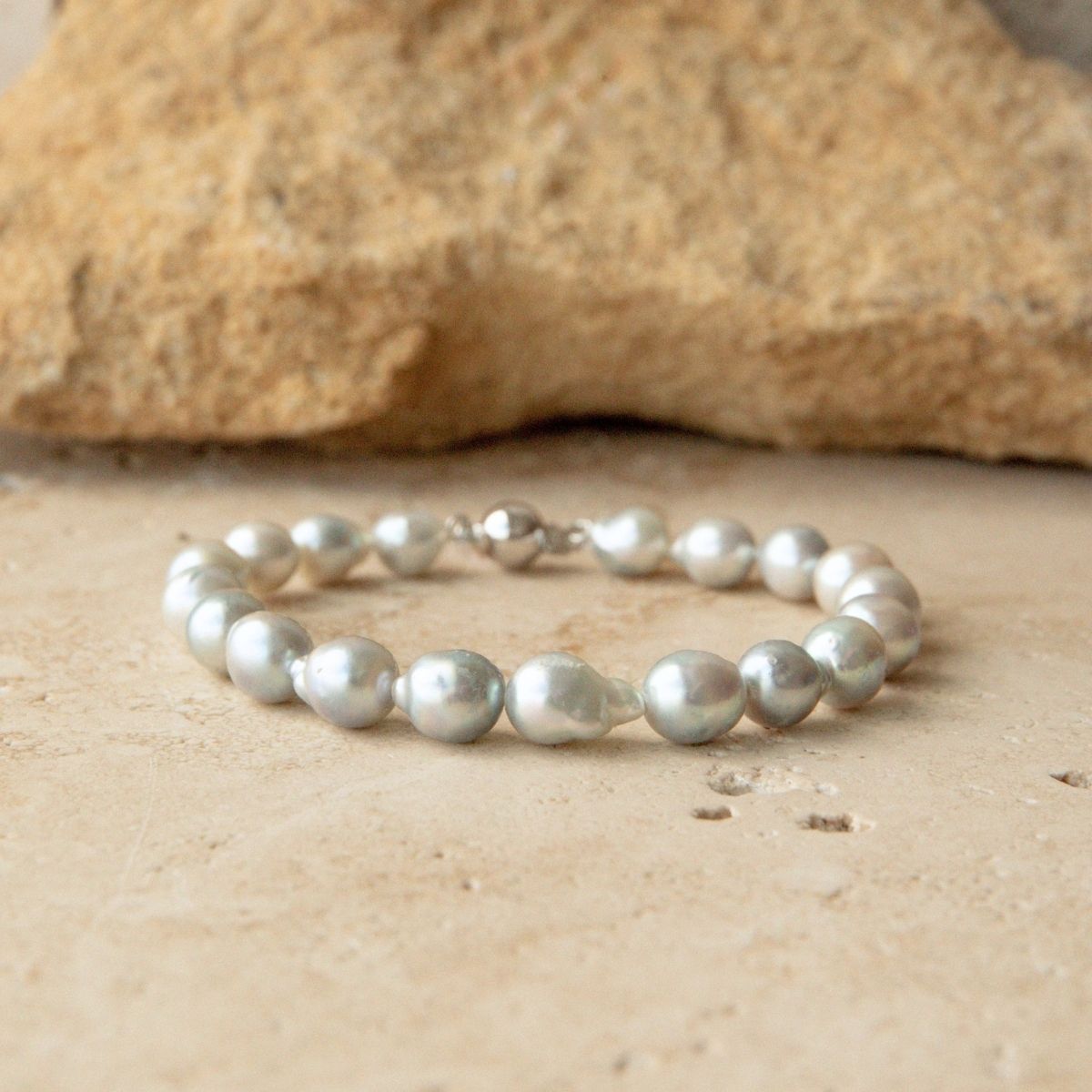 A bracelet made of irregular, lustrous silver toned Australian Akoya pearls with a solid gold clasp, displayed on a light beige stone surface with a larger rock in the background.