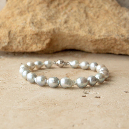A bracelet made of irregular, lustrous silver toned Australian Akoya pearls with a solid gold clasp, displayed on a light beige stone surface with a larger rock in the background.