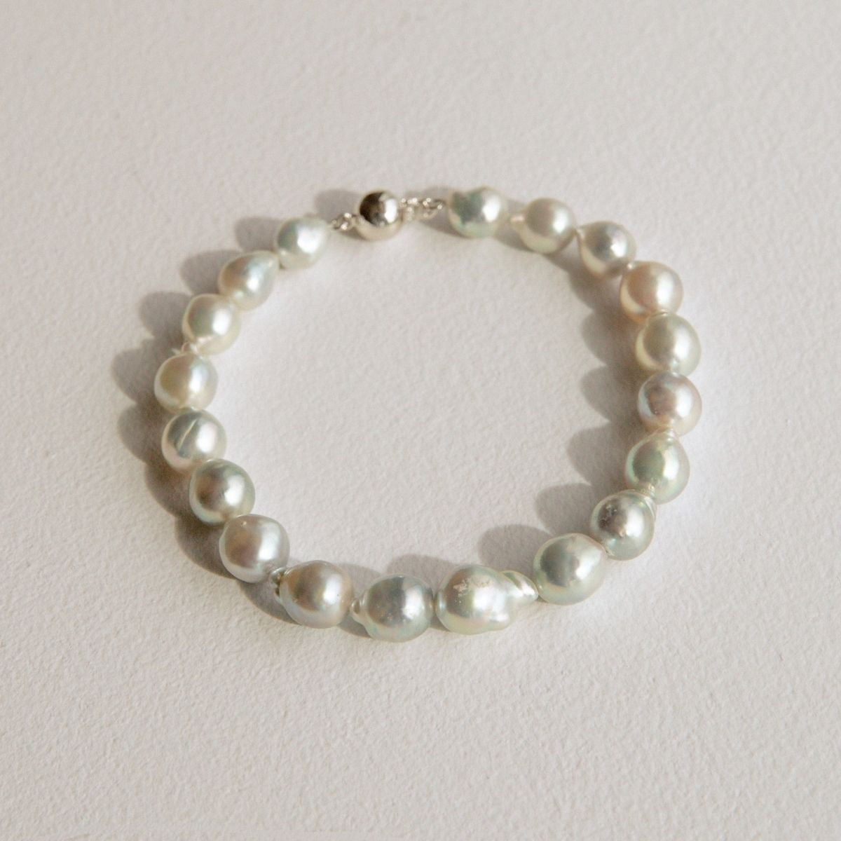A strand of irregular silver Australian Akoya pearls arranged in a circle with a small 18ct white gold clasp, photographed on a light beige textured surface.