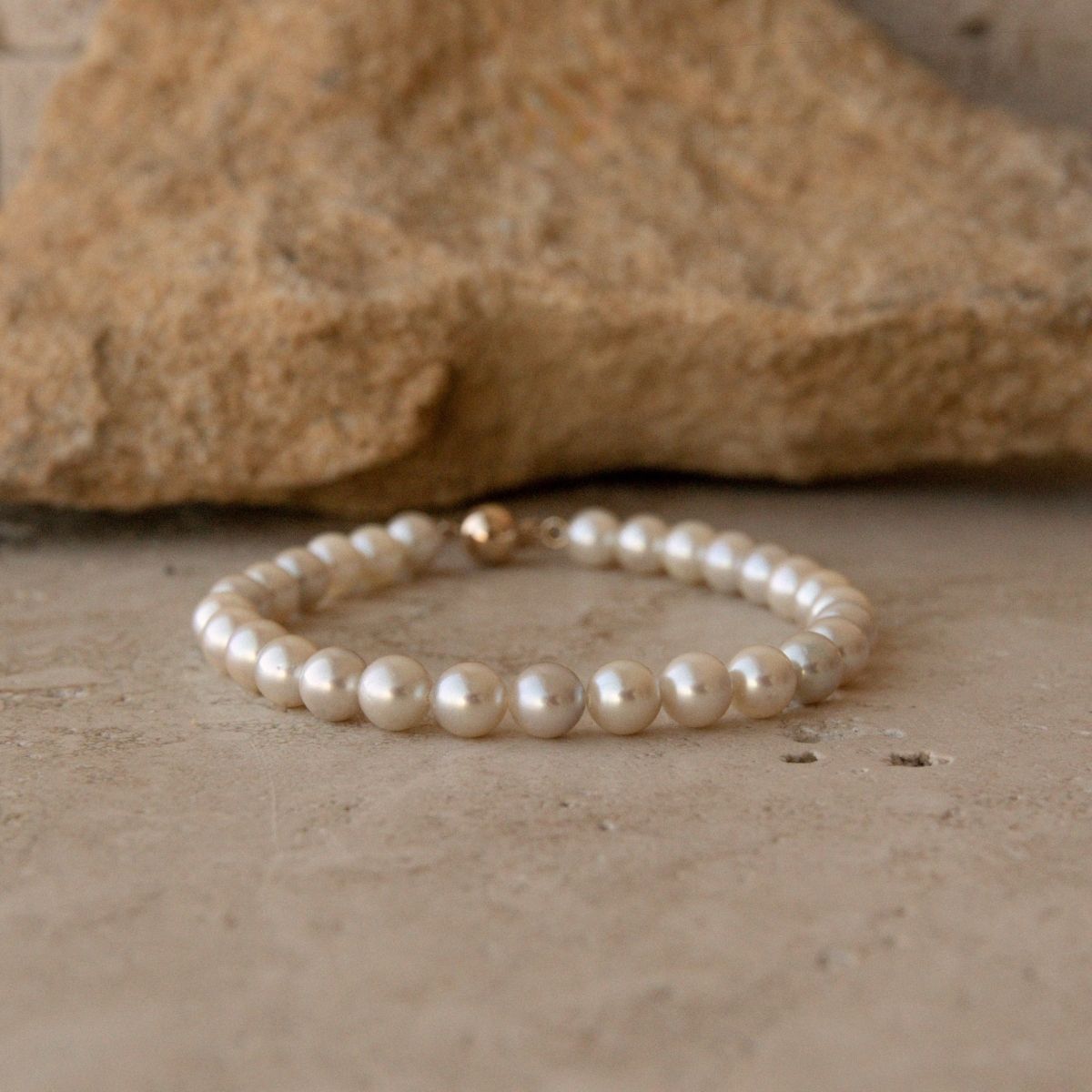 A pearl strand bracelet made of small Akoya pearls with a small 18ct yellow gold clasp, resting on a textured beige surface in front of a large tan rock.
