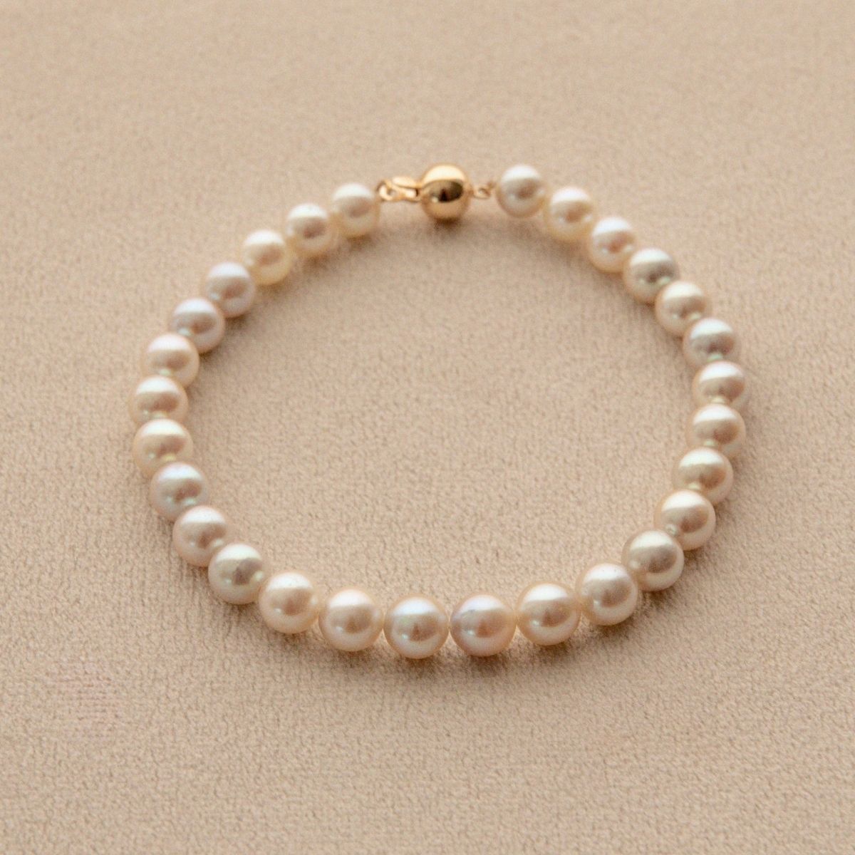 A single-strand pearl bracelet with a gold clasp, displayed on a textured beige surface.