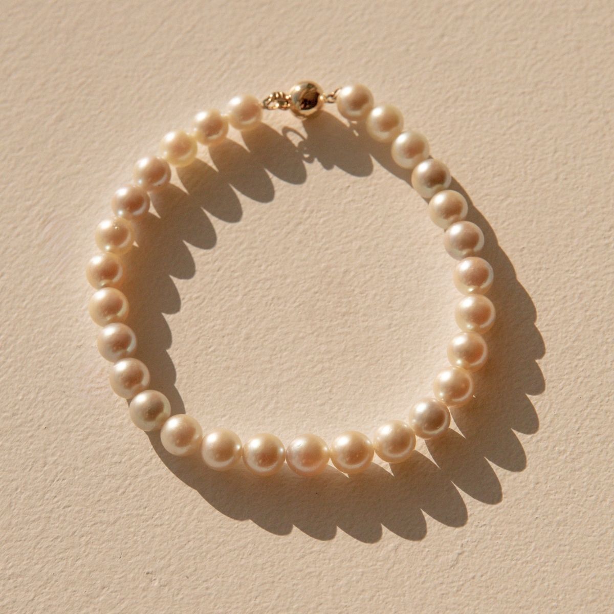 A single-strand pearl bracelet with a gold clasp, arranged in a circle on a textured beige surface with a strong diagonal shadow.