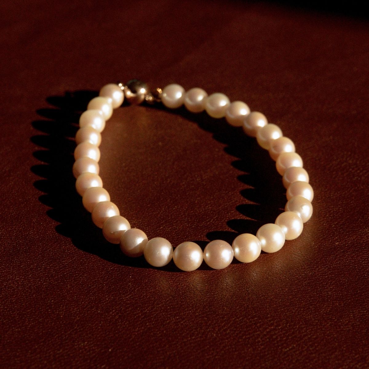 A single-strand pearl bracelet with a gold-toned clasp, resting on a dark brown textured surface under warm directional light.