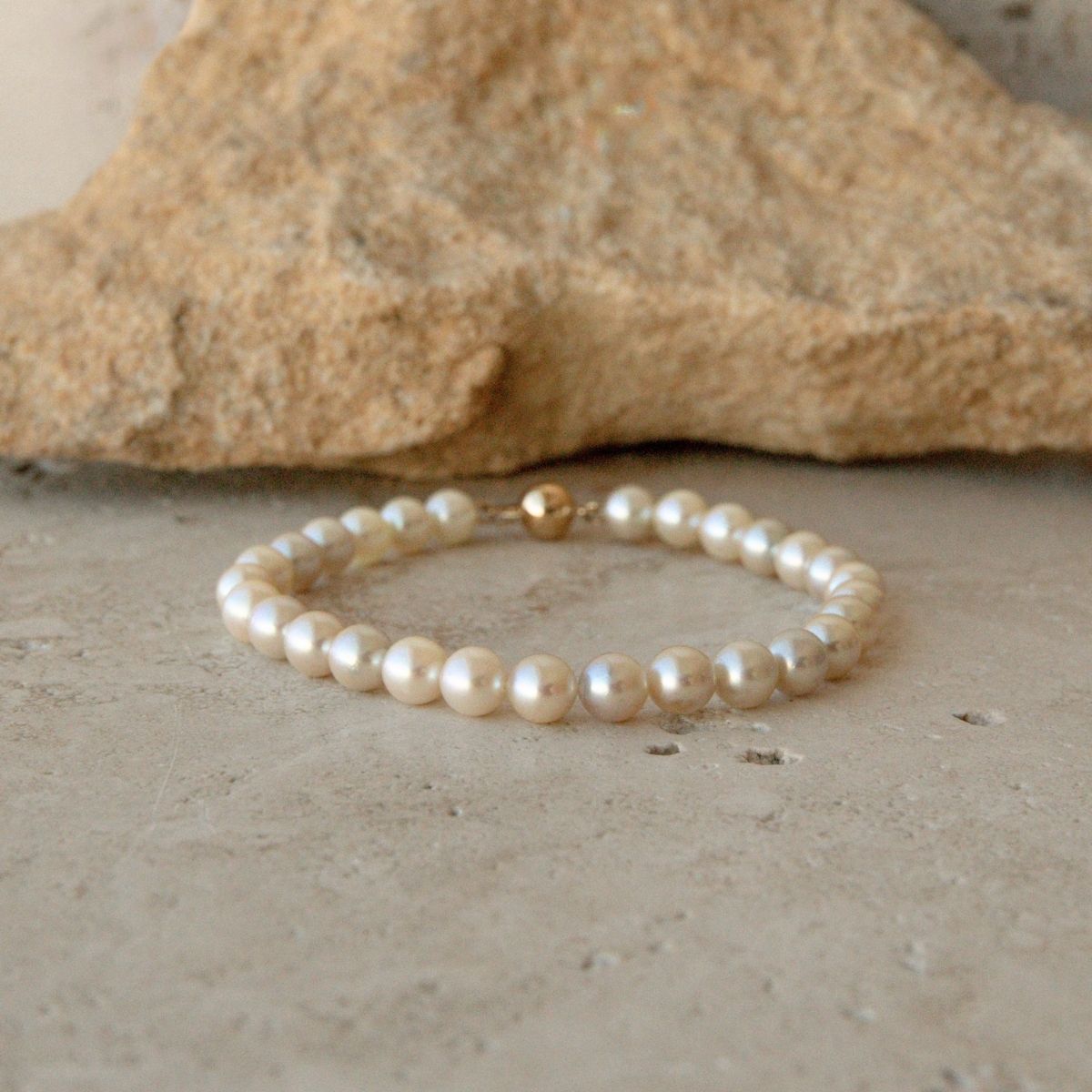 A pearl strand bracelet made of cream Australian Akoya pearls with a single gold ball clasp, resting on a textured stone surface with a larger rock in the background.