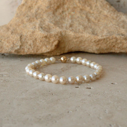A pearl strand bracelet made of cream Australian Akoya pearls with a single gold ball clasp, resting on a textured stone surface with a larger rock in the background.