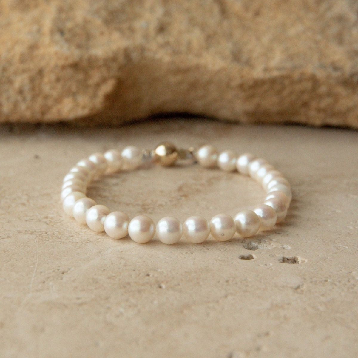 A pearl strand bracelet with a 9ct yellow gold clasp resting on a textured beige stone surface with a larger stone in the background.