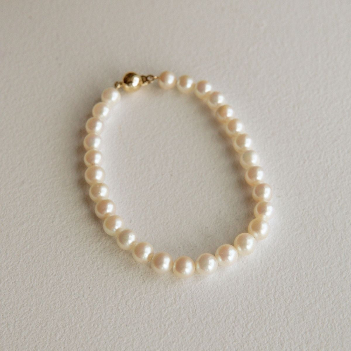 A single-strand Australian Akoya pearl bracelet with a gold clasp, arranged in an oval shape on a textured off-white background.