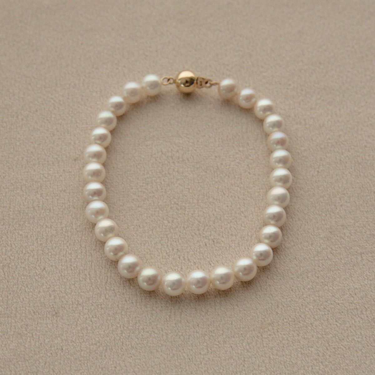A single-strand Akoya pearl bracelet with a gold clasp, displayed on a textured beige surface.