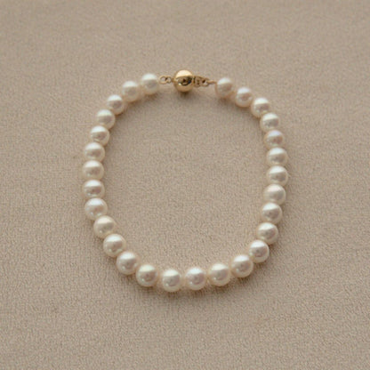 A single-strand Akoya pearl bracelet with a gold clasp, displayed on a textured beige surface.