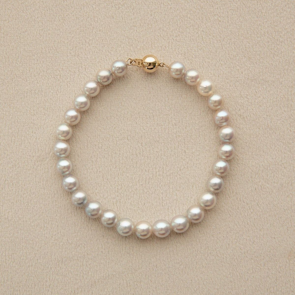 A classic Akoya pearl bracelet with a gold clasp, displayed on a textured beige surface.