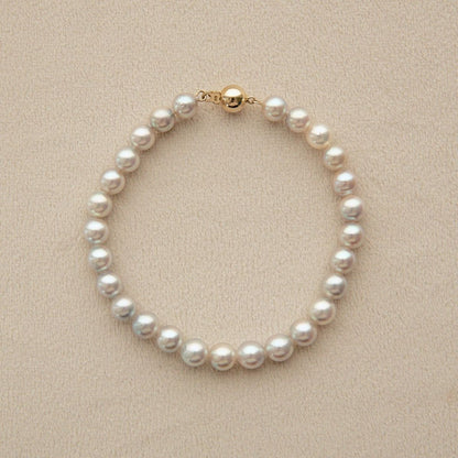 A classic Akoya pearl bracelet with a gold clasp, displayed on a textured beige surface.