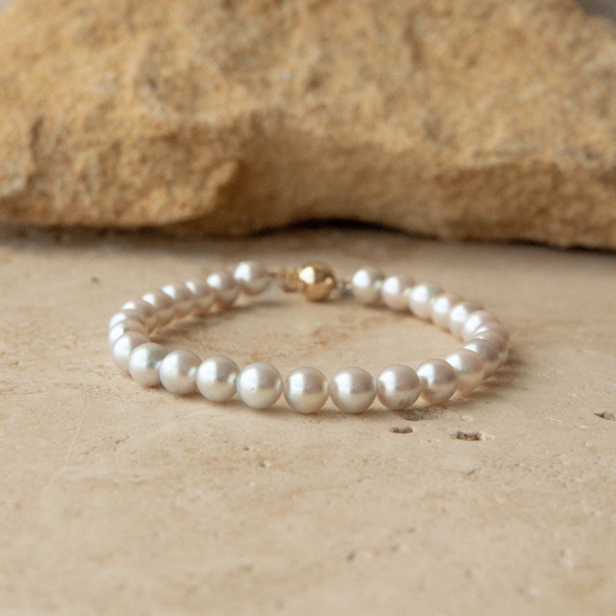A pearl strand bracelet made of round white Australian Akoya pearls with a small yellow gold ball clasp, resting on a beige stone surface with a larger rock in the background.