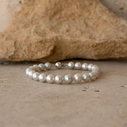 A pearl strand bracelet made of round, lustrous white Akoya pearls with a 9ct white gold ball clasp, photographed on a stone surface with a larger rock in the background.