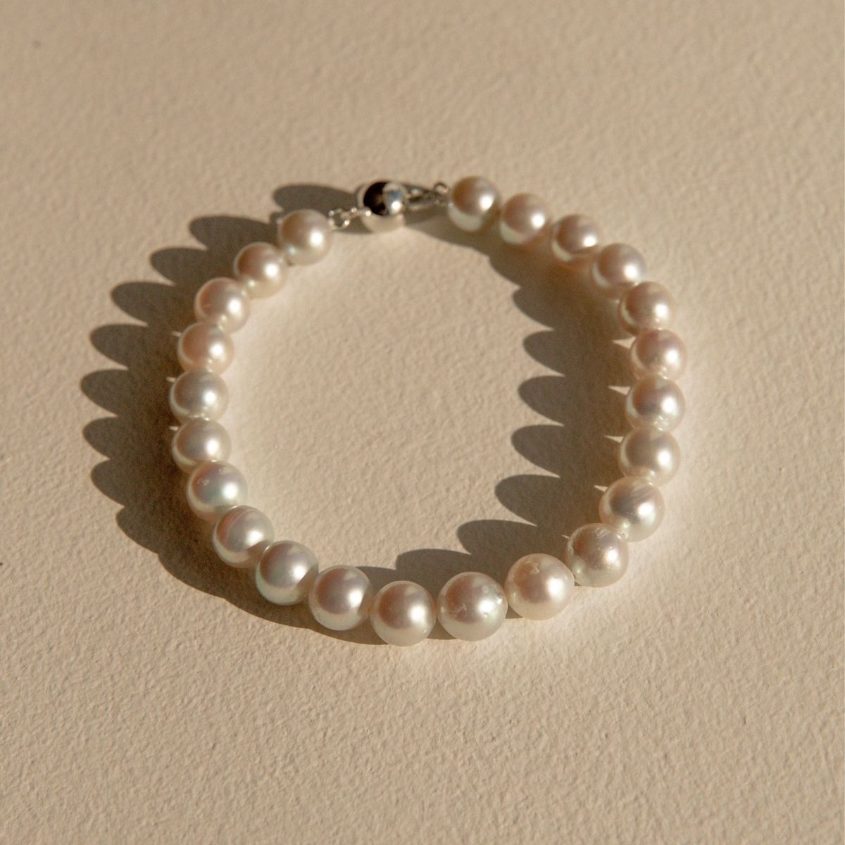 A single-strand pearl bracelet with a 9ct white gold ball clasp, displayed on a textured beige surface with a soft shadow.