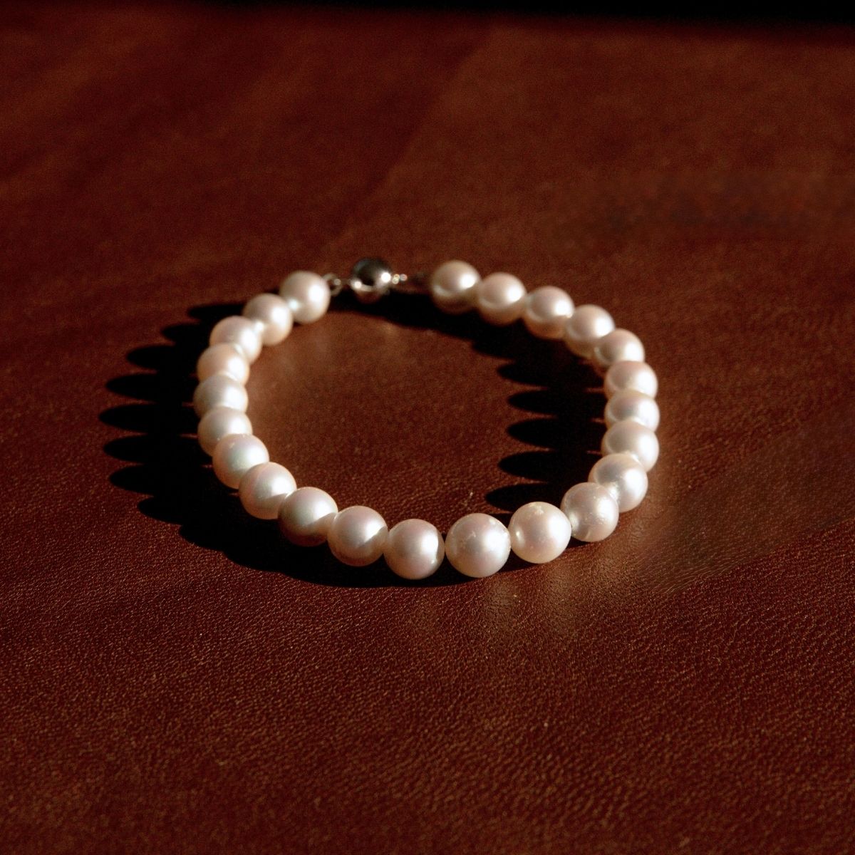 A pearl strand bracelet with a white gold ball clasp, resting on a dark brown textured leather surface.