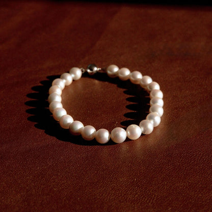 A pearl strand bracelet with a white gold ball clasp, resting on a dark brown textured leather surface.