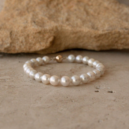 A pearl strand bracelet made of white Australian Akoya pearls with a small gold ball clasp, resting on a textured beige stone surface with a larger rock in the background.