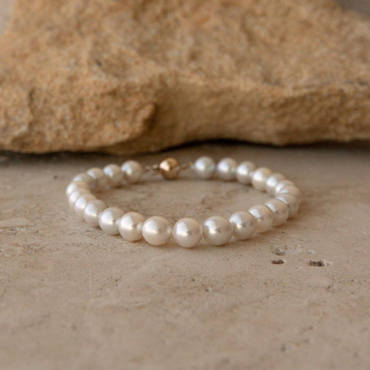A pearl strand bracelet made of white Australian Akoya pearls with a small gold ball clasp, resting on a textured beige stone surface with a larger rock in the background.