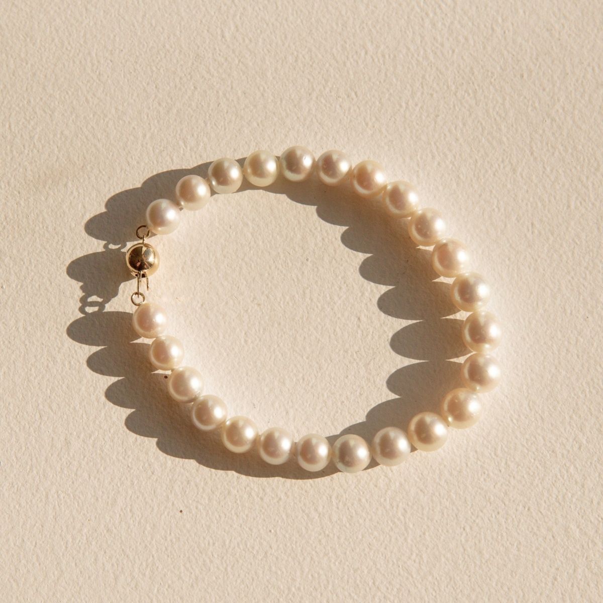 A single-strand Akoya pearl bracelet with a gold clasp, arranged in a circle on a textured beige background with soft shadows.