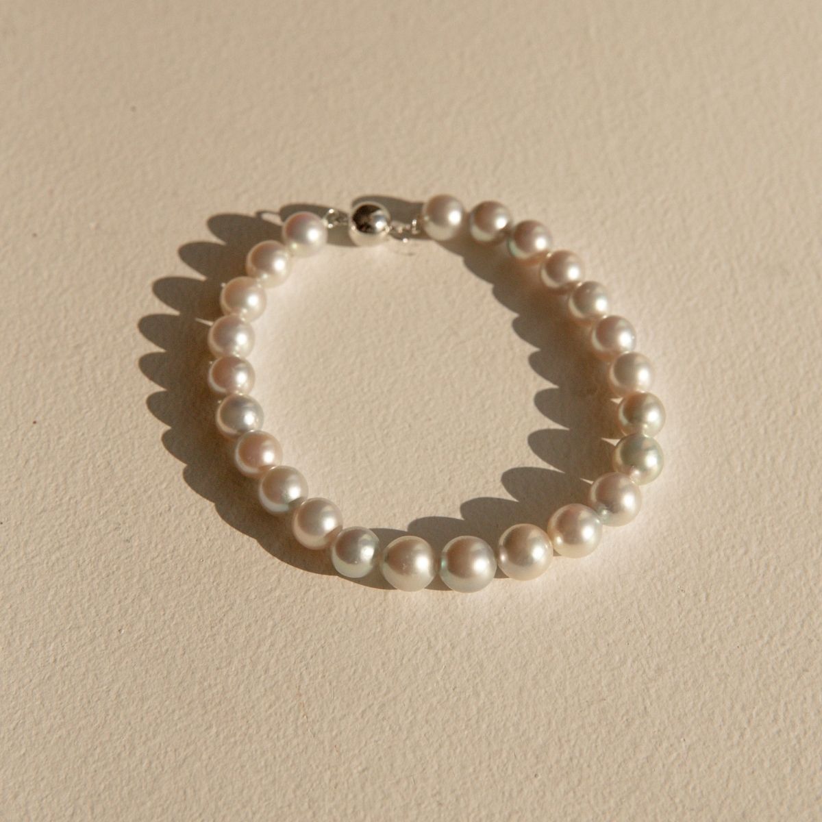 A classic pearl strand bracelet with a white gold ball clasp, displayed on a textured beige surface with soft shadows.