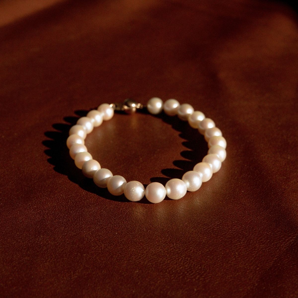 A pearl strand bracelet with an 18ct yellow gold clasp, resting on a dark brown textured leather surface.
