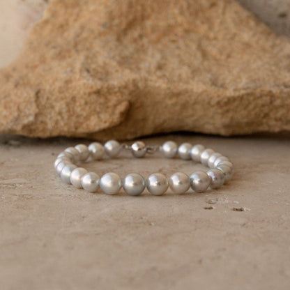 A bracelet made of round, lustrous silver toned Australian Akoya pearls, photographed on a stone surface with a larger rock in the background.