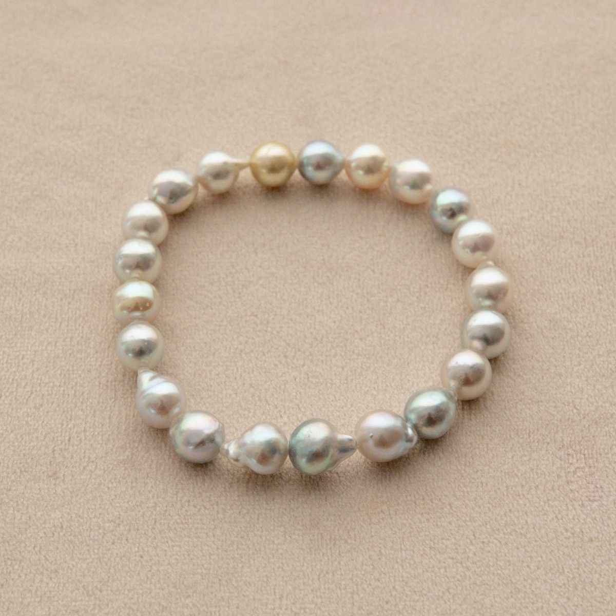 An elasticated pearl strand bracelet made of irregular baroque Akoya pearls in shades of white, cream, and silver, displayed on a textured beige background.