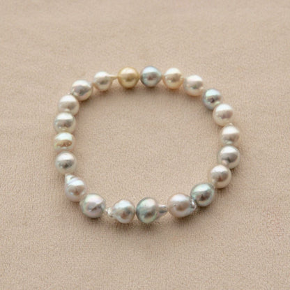 An elasticated pearl strand bracelet made of irregular baroque Akoya pearls in shades of white, cream, and silver, displayed on a textured beige background.