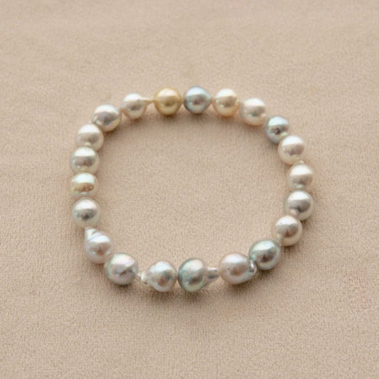 An elasticated pearl strand bracelet made of irregular baroque Akoya pearls in shades of white, cream, and silver, displayed on a textured beige background.