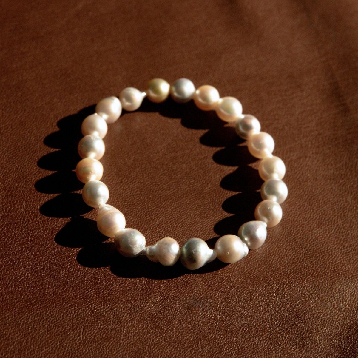 A pearl strand bracelet made of irregular, baroque Akoya pearls arranged in a circle on a textured brown leather background.