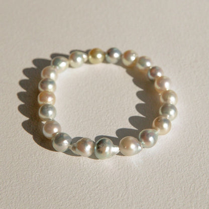 An Australian Akoya pearl strand bracelet made of irregular, lustrous pearls in soft champagne, cream, and silver tones, arranged in a circle on a textured off-white surface with a sharp shadow.