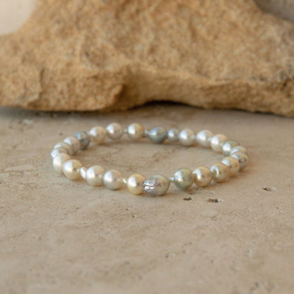 An elasticated pearl strand bracelet made of irregular Australian Akoya pearls in cream, white, and iridescent silver tones, resting on a textured beige stone surface with a larger rock blurred in the background.