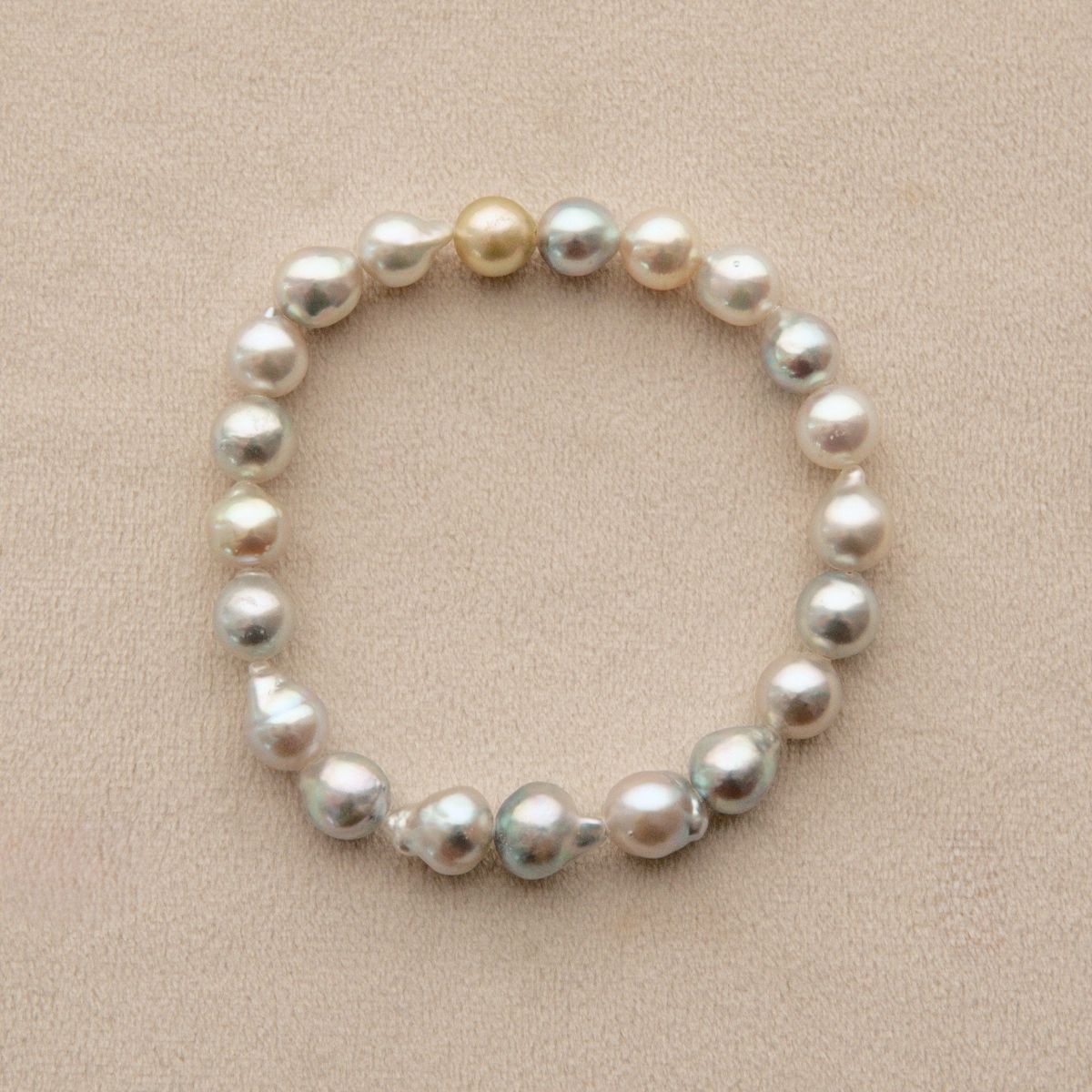 A beaded stretch bracelet made of irregular freshwater pearls in white, cream, and light gold tones, displayed on a textured beige fabric background.