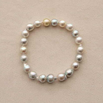 A beaded stretch bracelet made of irregular freshwater pearls in white, cream, and light gold tones, displayed on a textured beige fabric background.