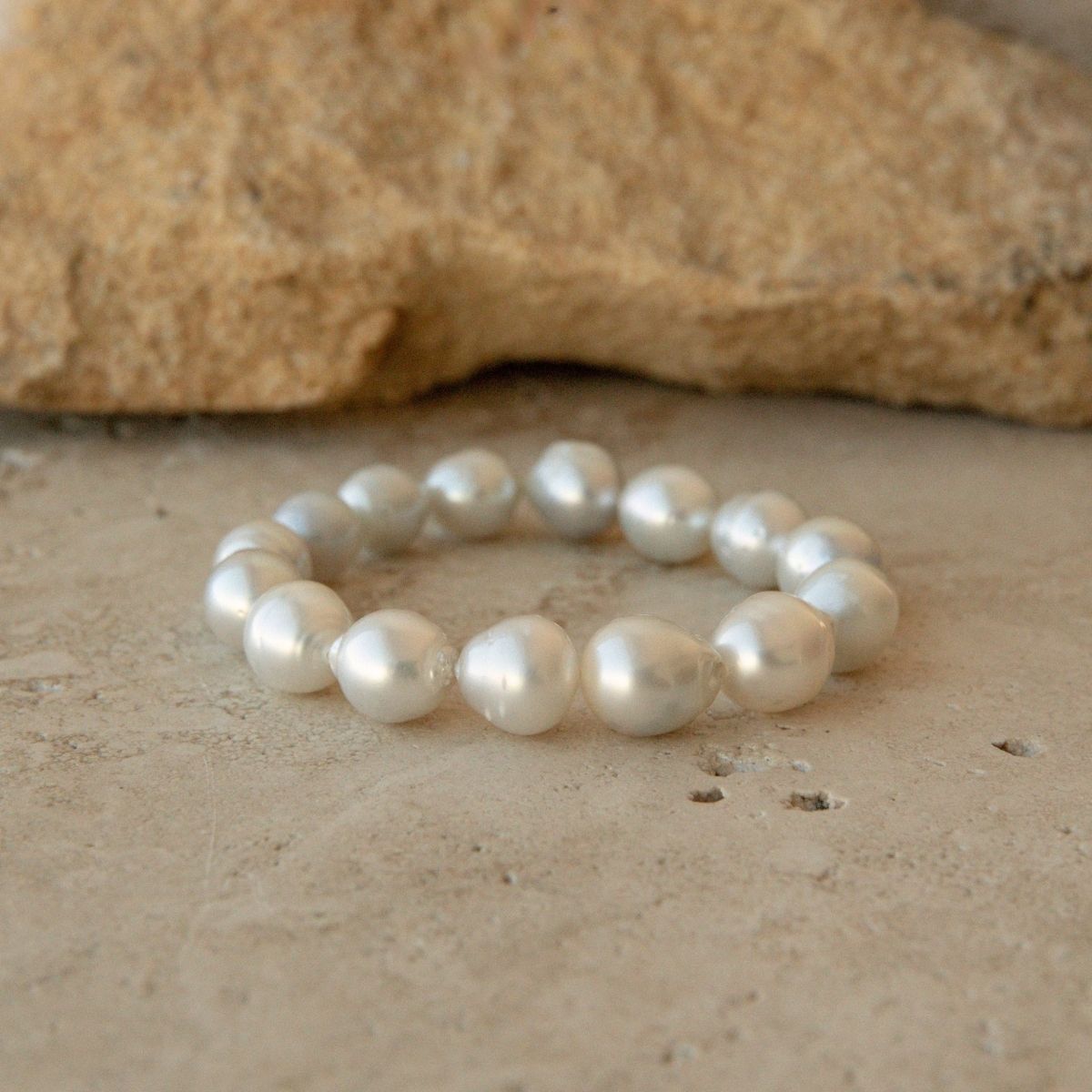 A pearl strand bracelet made of baroque Australian South Sea pearls, displayed on a textured beige stone surface with a larger tan rock in the background.