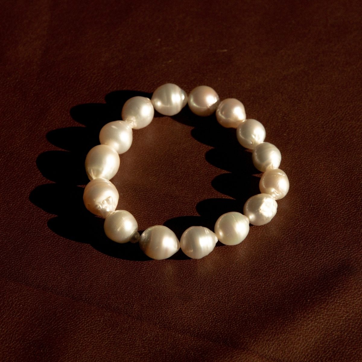 A barqoue pearl strand bracelet made of irregular Australian South Sea pearls, arranged in a circle on a dark brown textured leather background.