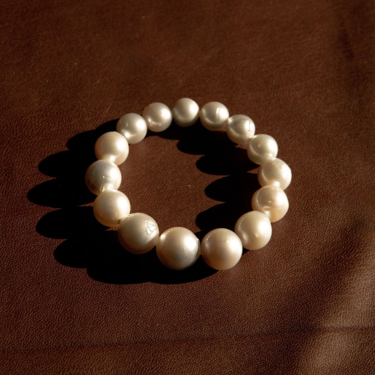A strand bracelet made of large, lustrous white Australian South pearls, arranged in a circle on a textured brown leather background.
