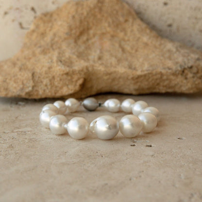 A baroque pearl strand bracelet with a single 9ct white gold ball clasp, displayed on a textured beige stone surface in front of a larger rock.