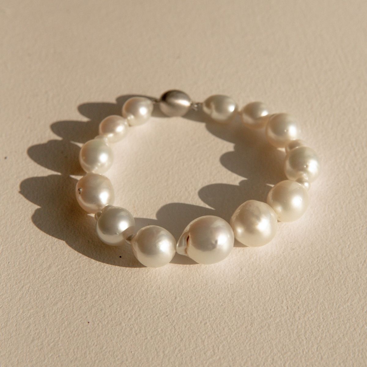 A baroque pearl bracelet with irregular white Australian South Sea pearls and a 9ct white gold clasp, photographed on a textured beige surface with a soft shadow.