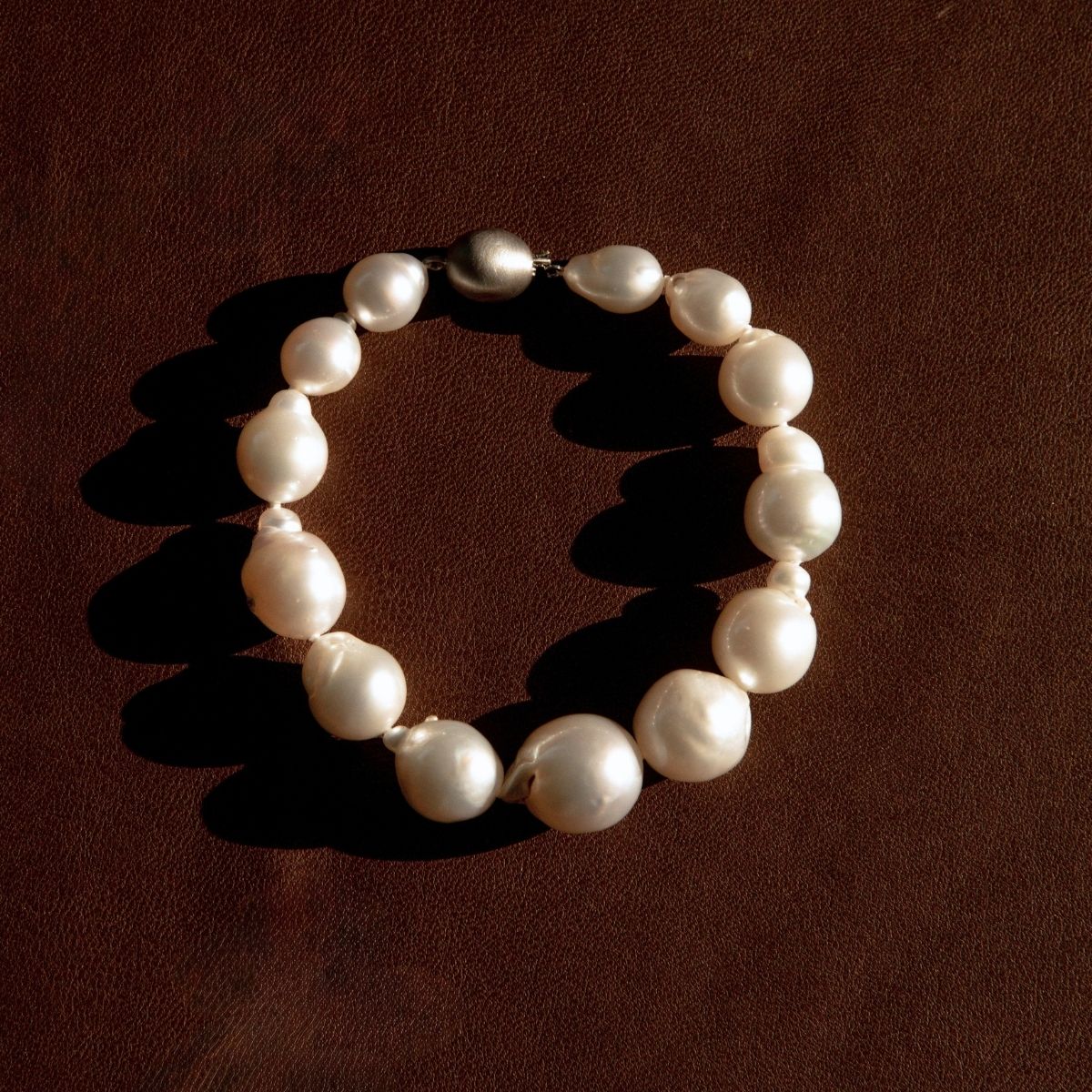 A pearl strand bracelet made of irregular white baroque pearls arranged in a circle on a dark brown textured background.