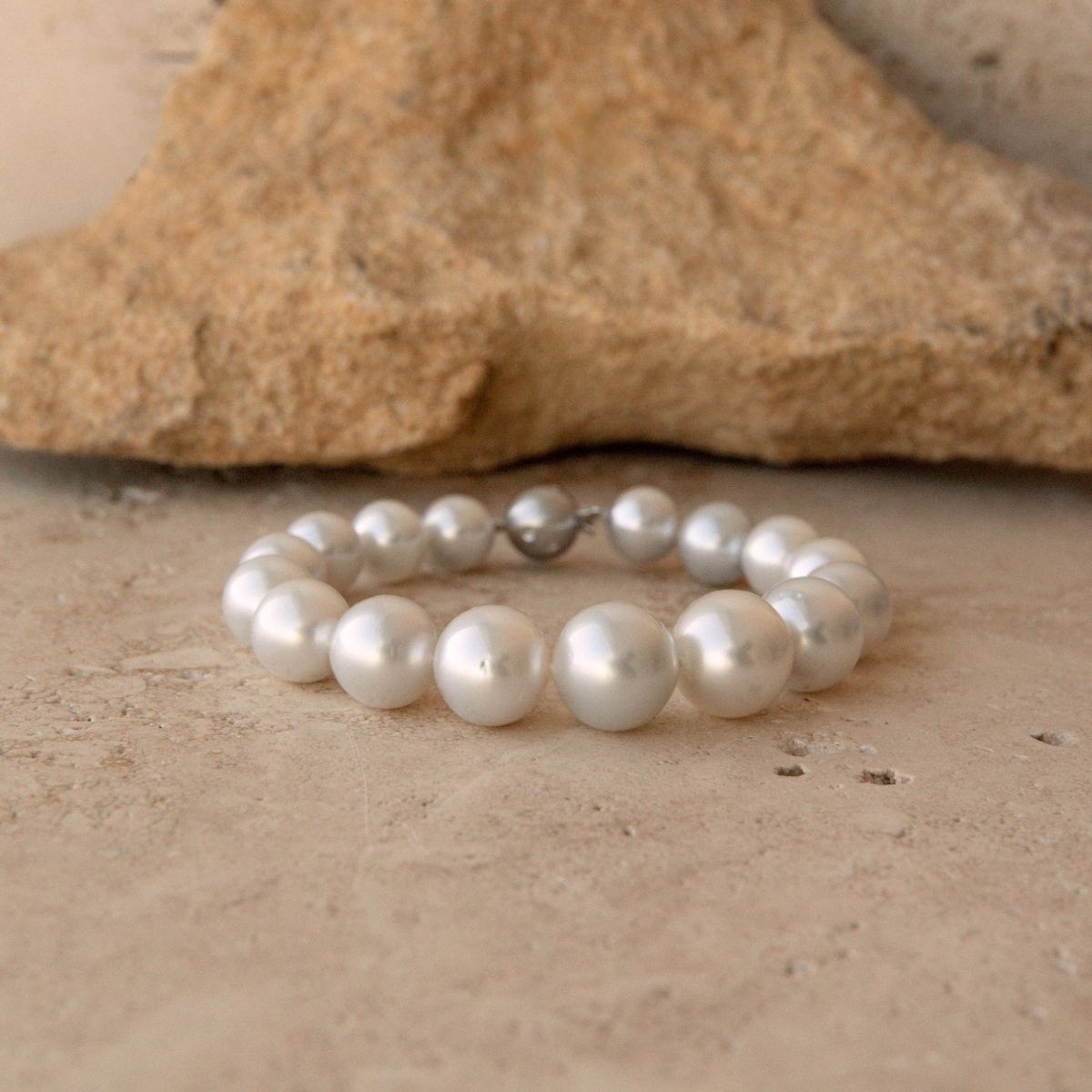 A pearl strand bracelet made of large, lustrous white Saltwater pearls with a small 9ct white gold clasp, displayed on a textured beige stone surface with a larger rock in the background.
