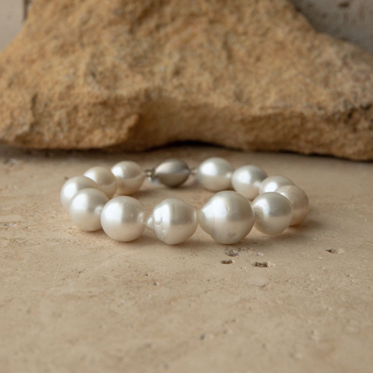 A bracelet made of large, lustrous white Australian South Sea pearls with a small 9ct white gold clasp, displayed on a textured beige stone surface with a larger rock in the background.