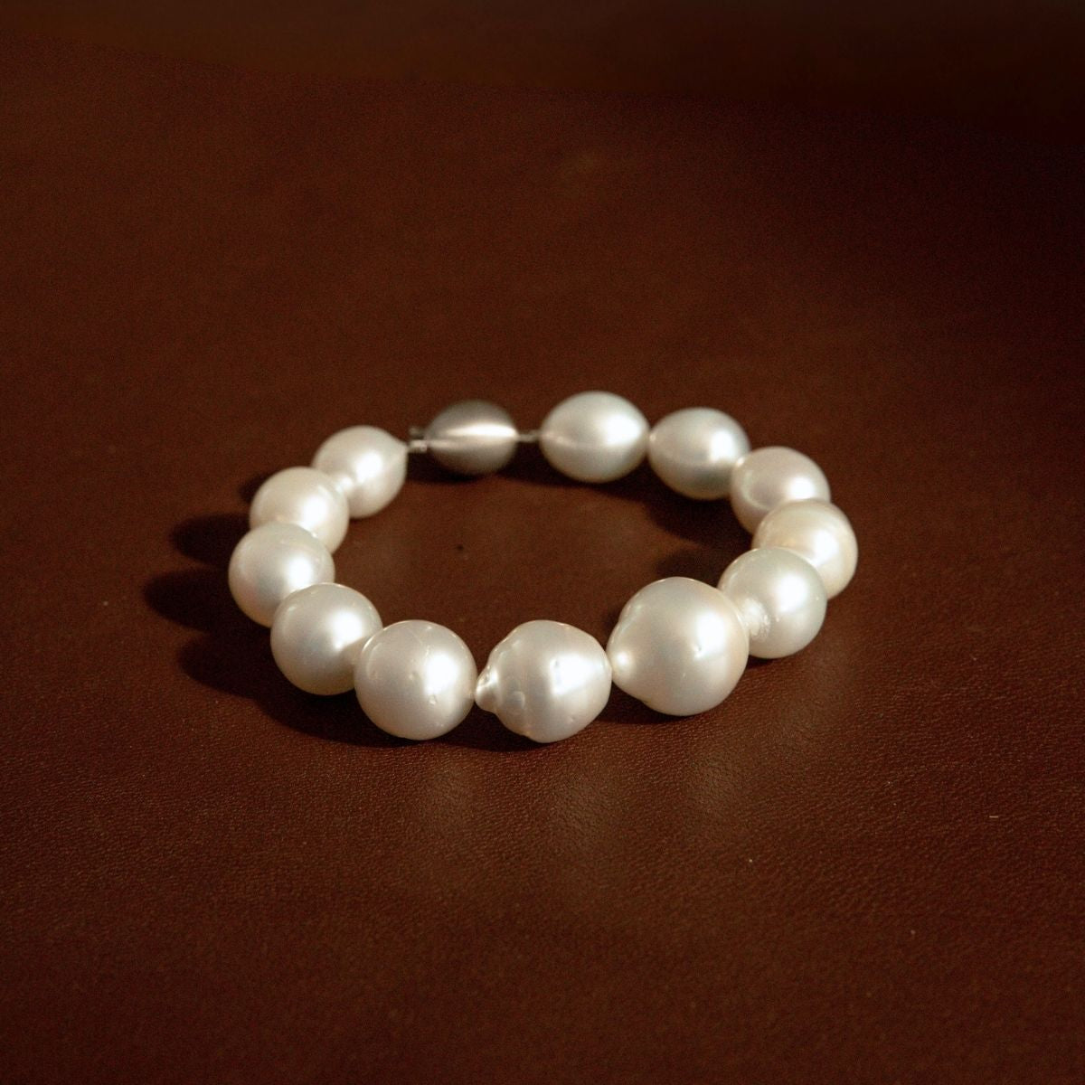 An Australian South Sea pearl strand bracelet made of irregular white Saltwater pearls and one metallic 9ct white gold bead, displayed on a dark brown leather surface.